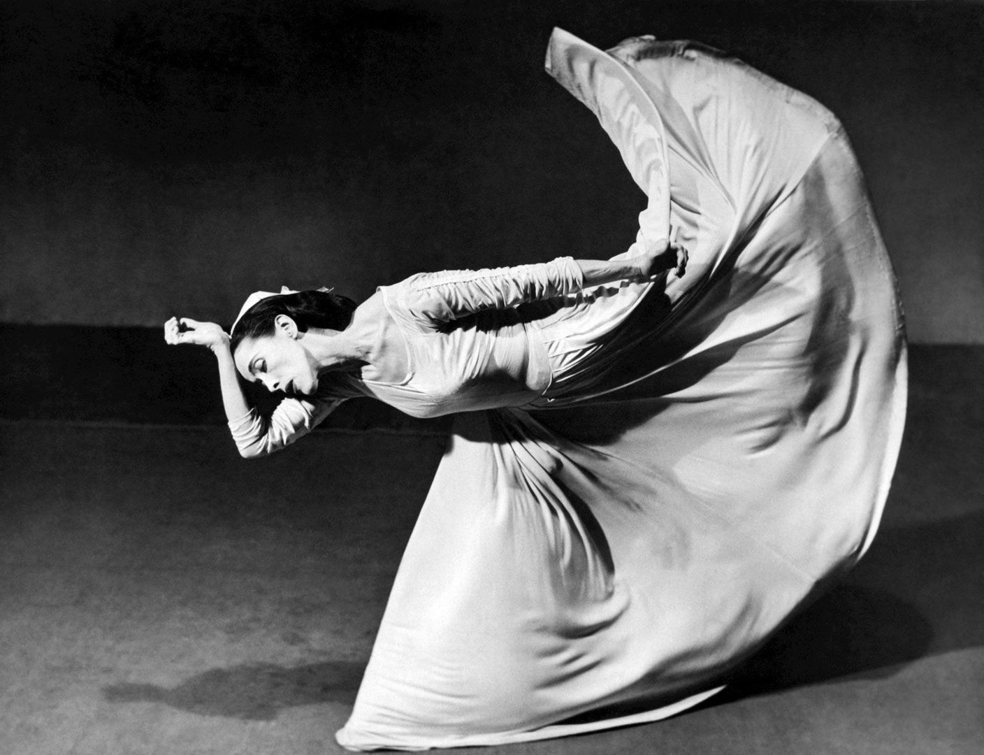 A black-and-white photo of a woman dancing in a flowing dress. Her torso is horizontal to the ground, and one of her legs, covered by the long dress, is kicked up behind her.